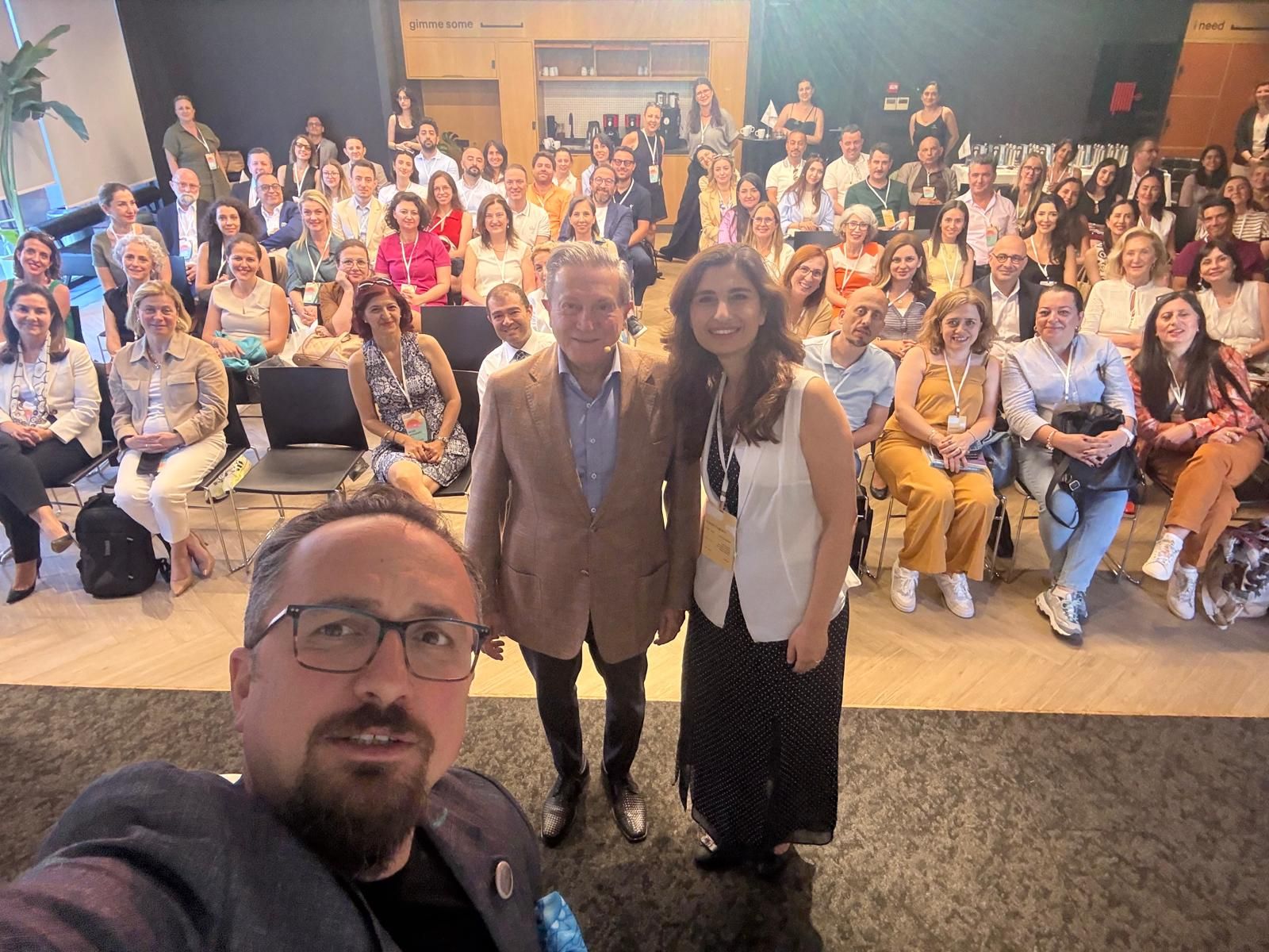 A group photo featuring a diverse audience at an event, with three individuals in the foreground posing for a selfie.